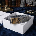 A Dior belt with a gold-tone 'DIOR' buckle and a dark monogram patterned strap, presented inside an open white Dior gift box on a dark blue velvet surface with bookshelves in the background.