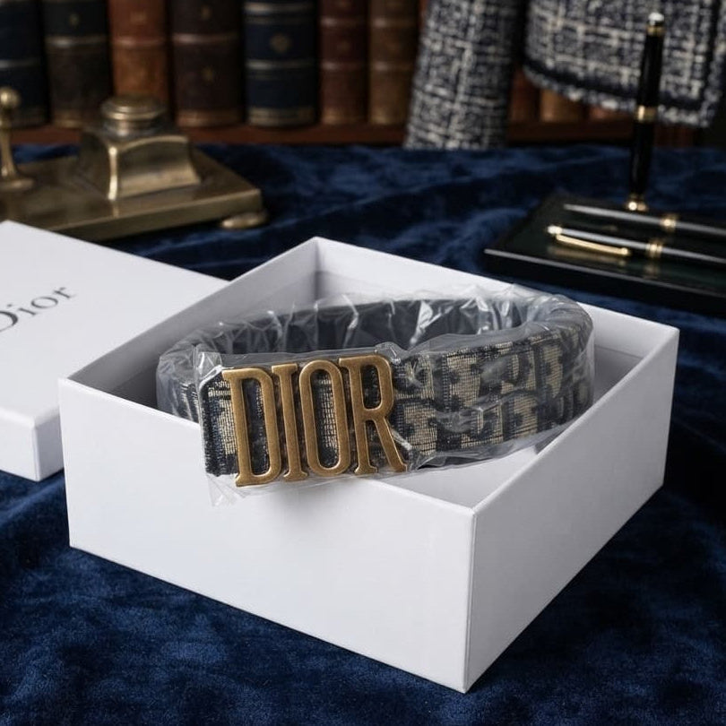 A Dior belt with a gold-tone 'DIOR' buckle and a dark monogram patterned strap, presented inside an open white Dior gift box on a dark blue velvet surface with bookshelves in the background.