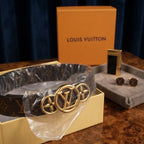 A Louis Vuitton monogram belt with a large gold-tone LV logo buckle, shown in its packaging with an orange Louis Vuitton box and a gray jewelry tray in the background.